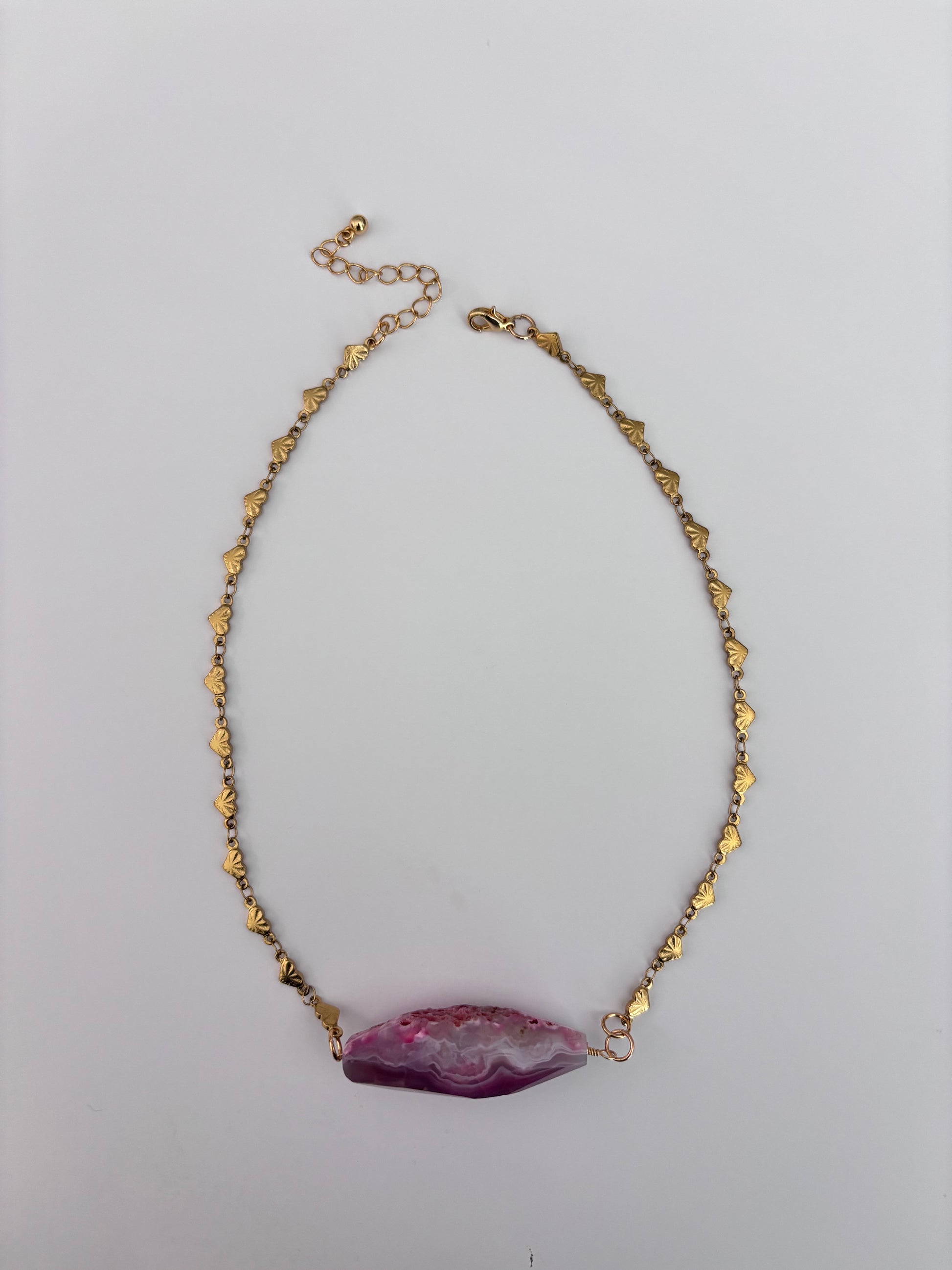 A gold chain necklace with small metallic leaf-shaped charms and a large, polished purple agate geode pendant, displayed on a light gray background.