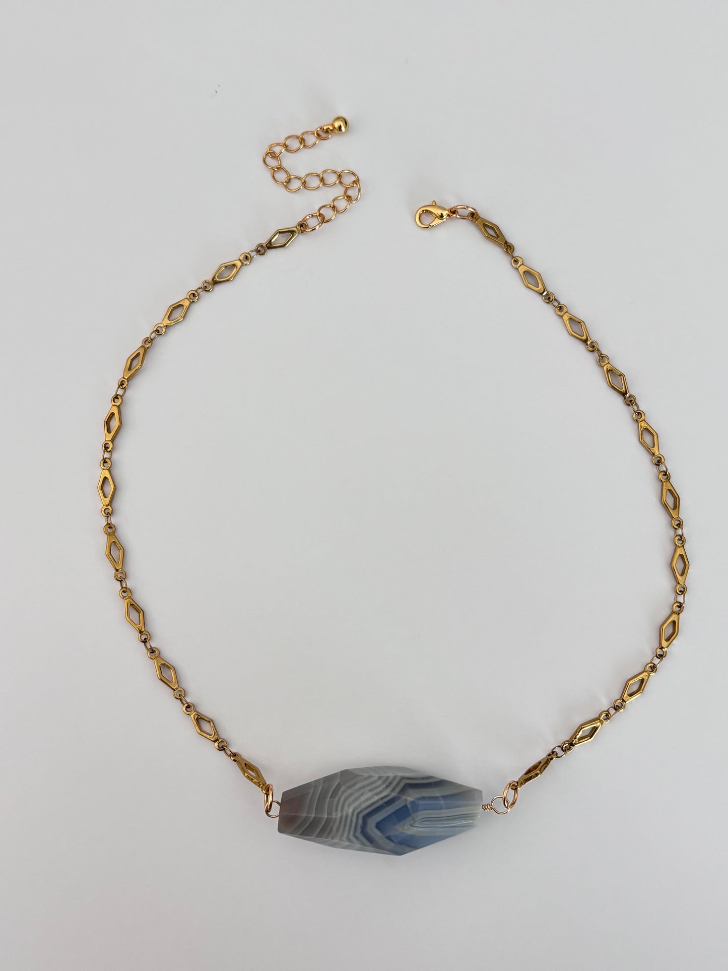 A gold-toned chain necklace with a faceted blue and gray agate stone pendant, displayed on a light gray background.