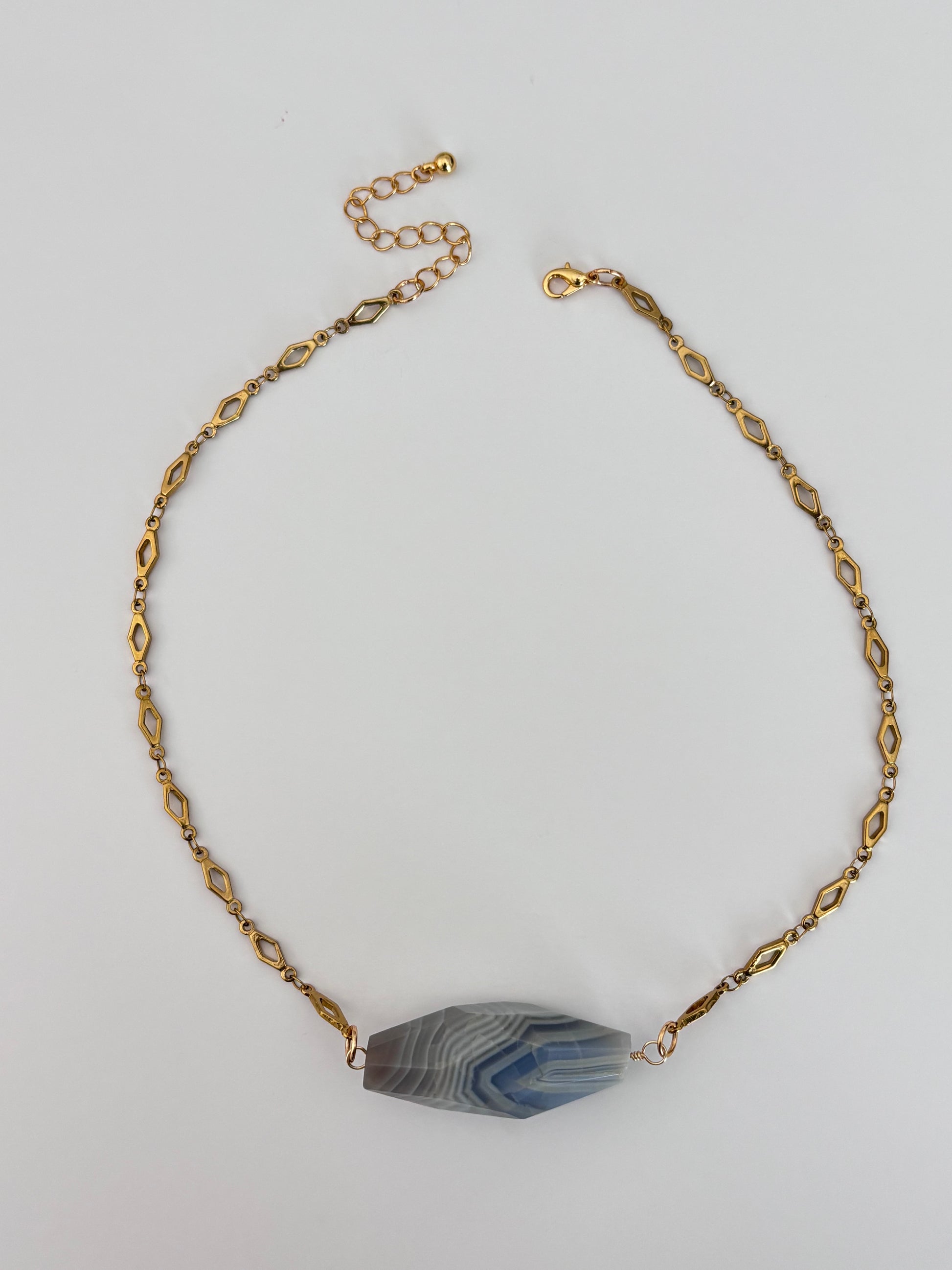 A gold-toned chain necklace with a faceted blue and gray agate stone pendant, displayed on a light gray background.